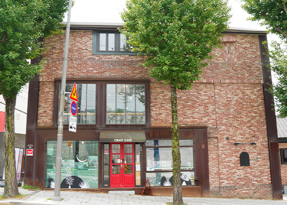 붉은 벽돌로 된 3층 건물 1층에 빨간 출입문이 있는 카페와 ‘CRAFT CAFE’ 간판이 보이는 거리 풍경