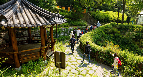 KCU 걷기대회 두번째 이미지, 전통 기와지붕 정자가 있는 푸른 공원 산책로를 따라 걸어가고 있는 사람들의 뒷모습