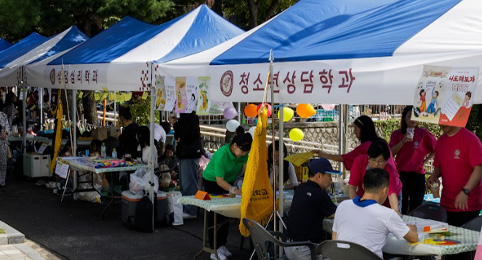 KCU 가을축제 첫번째 이미지, 야외 광장에서 무대를 향해 양손을 위로 들고 환호하고 있는 사람들의 뒷모습과 무대 위 사람들의 모습